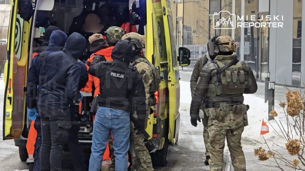 Grupa funkcjonariuszy w mundurach, w tym policjanci i ratownicy medyczni, pomaga osobie wsiadającej do karetki na zaśnieżonej ulicy.