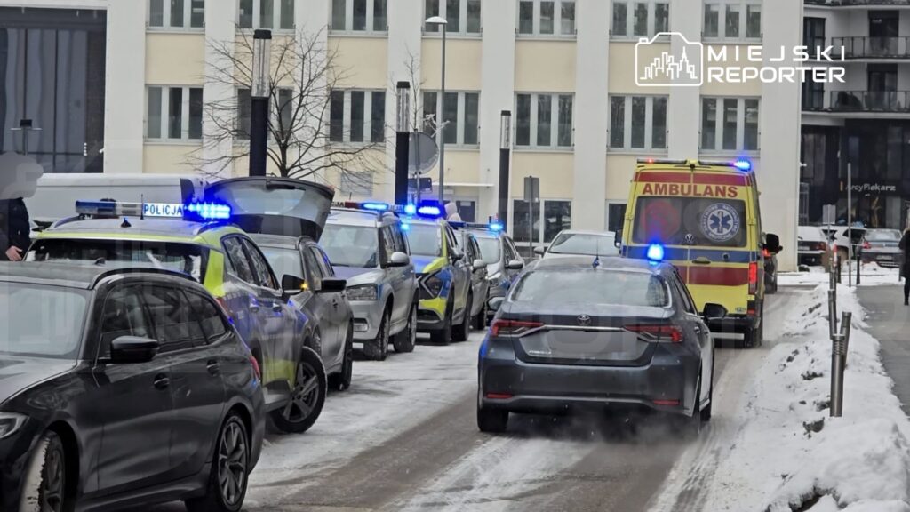 Grupa pojazdów policyjnych i karetka na zaśnieżonej ulicy, w tle nowoczesne budynki.