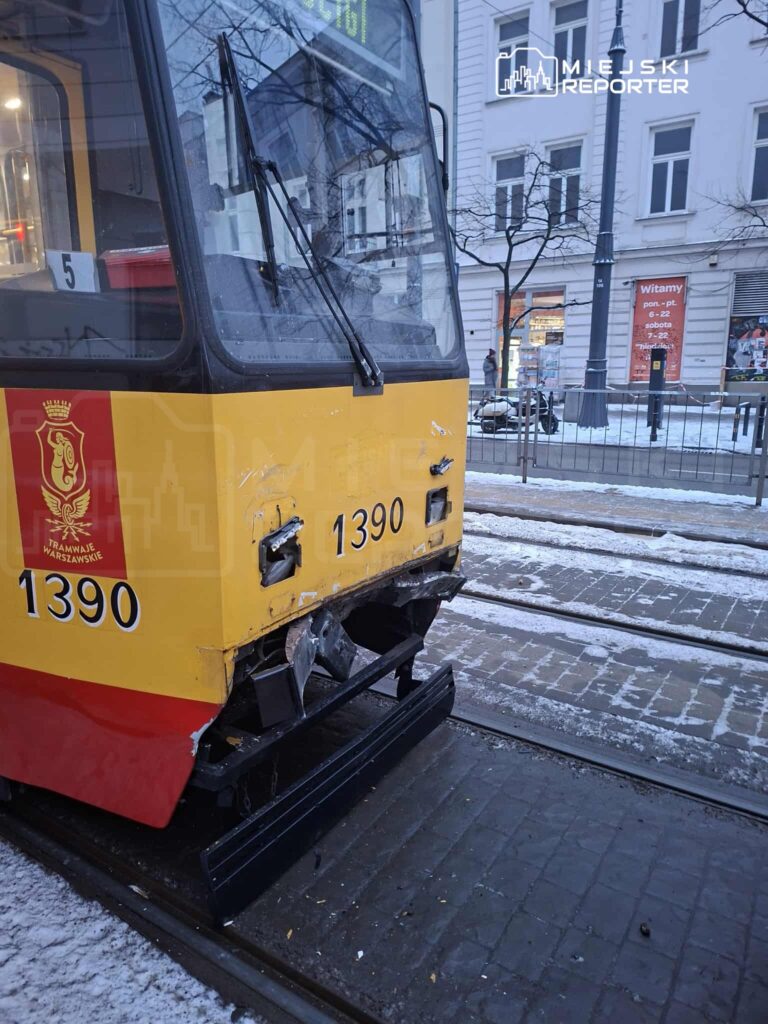Tramwaj z numerem 1390 z uszkodzeniami na przedniej części stoi na zimowej ulicy, obok przystanku.