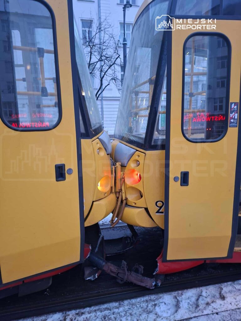 Dwa żółte tramwaje zderzone na torach, z widocznymi uszkodzeniami i włączonymi światłami, w miejskim otoczeniu.