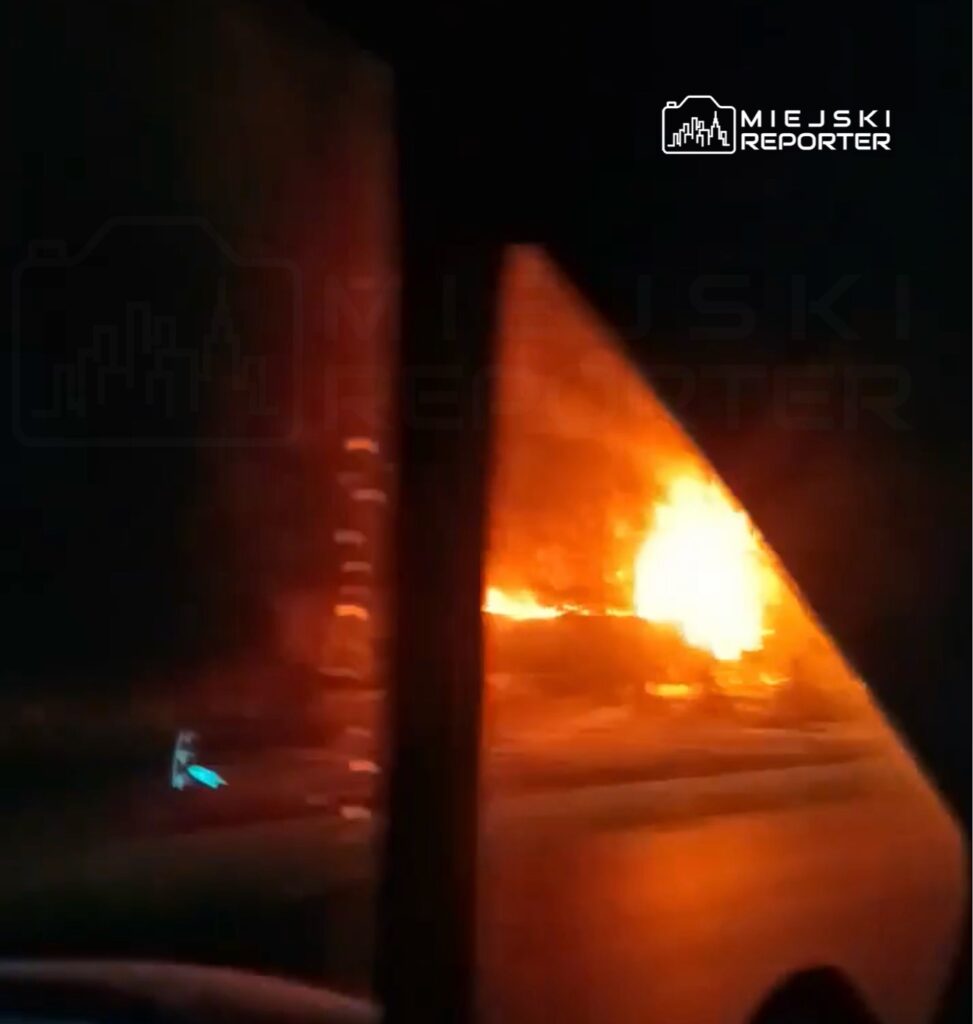 W nocy widać intensywny ogień wydobywający się z płonącego obiektu w pobliżu drogi.