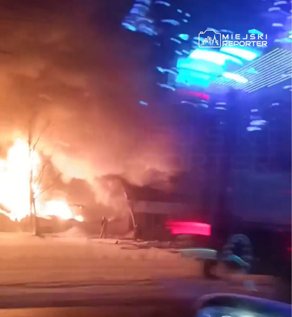 Pożar budynku z intensywnymi płomieniami i dymem, w tle widoczne niebieskie światła pojazdu służb ratunkowych.