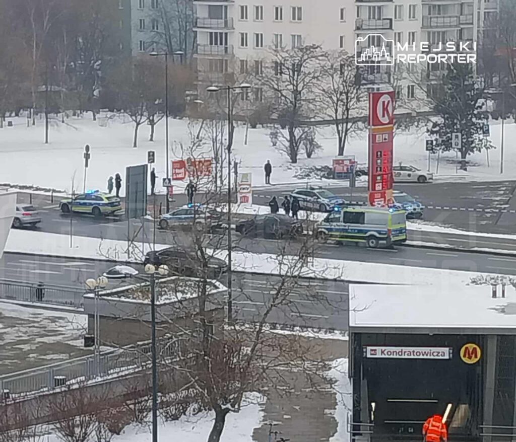 Grupa policjantów i karetka na zablokowanej ulicy, obok stacji metra w zimowym otoczeniu.
