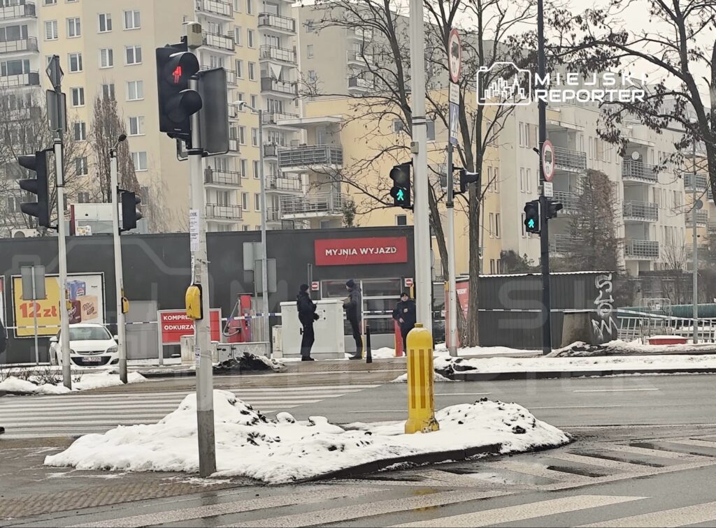 Na przejściu dla pieszych stoją dwaj mężczyźni w mundurach, a obok nich widoczny jest czerwony sygnalizator świetlny. W tle znajduje się myjnia oraz budynki mieszkalne. Na chodniku leży śnieg.