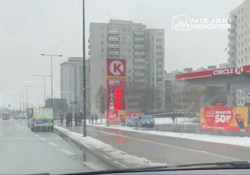 Na jezdni zatrzymany ambulans, obok grupka ludzi i dwa radiowozy, w tle nowoczesne budynki oraz stacja benzynowa.