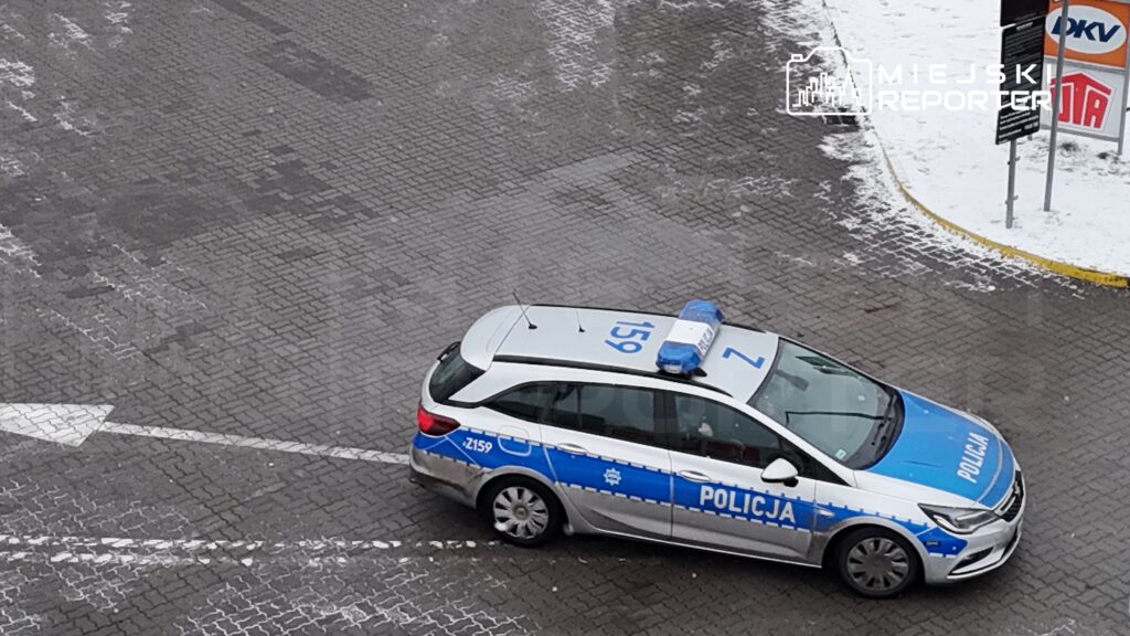 Policyjny radiowóz z niebieskimi światłami porusza się po wybrukowanej drodze w miejskim otoczeniu.
