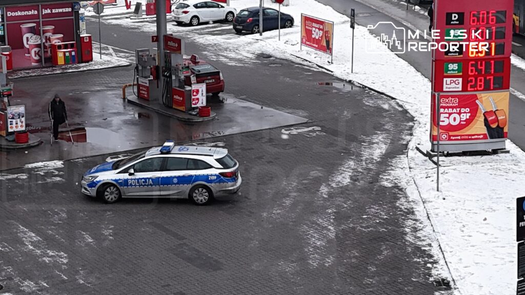 Policyjny radiowóz z niebieskimi i białymi oznaczeniami stoi na stacji paliw, a mężczyzna z laską przechodzi obok.