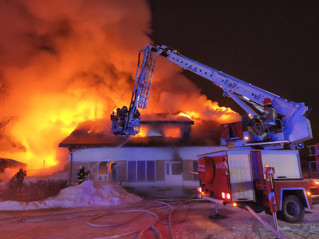 Strażacy gaszą pożar w nocy za pomocą drabiny mechanicznej, otaczająca budynek w ogniu i dymie.