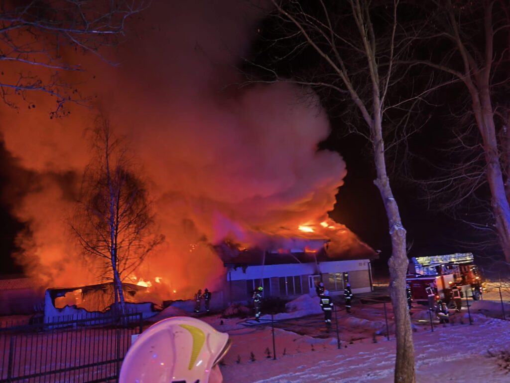 Płonący budynek w nocy, otoczony przez strażaków gaszących ogień, z gęstym dymem unoszącym się w powietrzu.