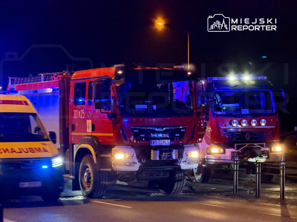 Dwa samochody strażackie z włączonymi sygnalizacjami świetlnymi stoją na drodze w nocy, obok karetki pogotowia.