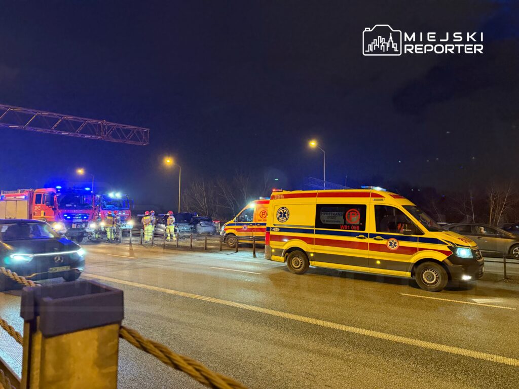 Na drodze nocą zderzenie samochodów, obok pracują strażacy i zespół medyczny przy ambulansie.