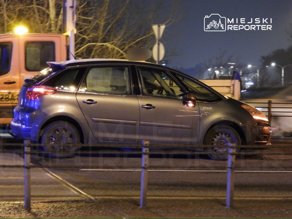 Samochód osobowy w ciemnoszarym kolorze zaparkowany na poboczu ulicy w nocy, obok pomarańczowego pojazdu.