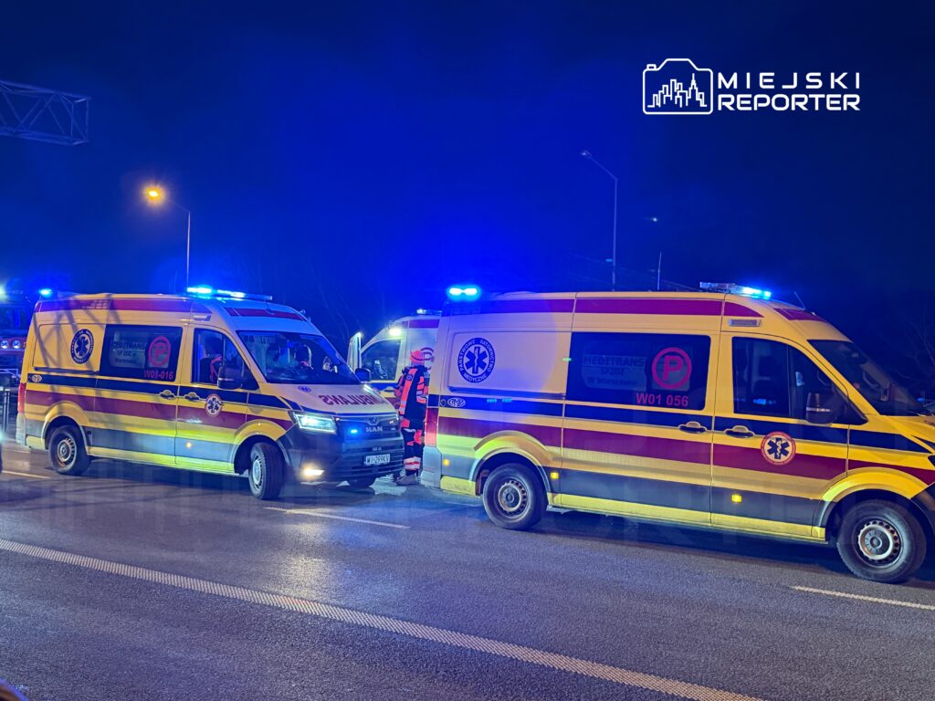 Dwa karetki pogotowia z włączonymi sygnalizacjami świetlnymi na drodze w nocy, otoczone przez personel medyczny.
