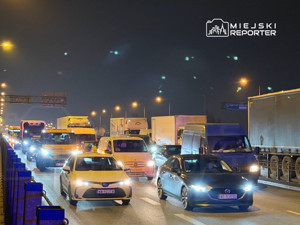 Ruch samochodowy na zatłoczonej drodze nocą, z widocznymi światłami pojazdów i znakami drogowymi.