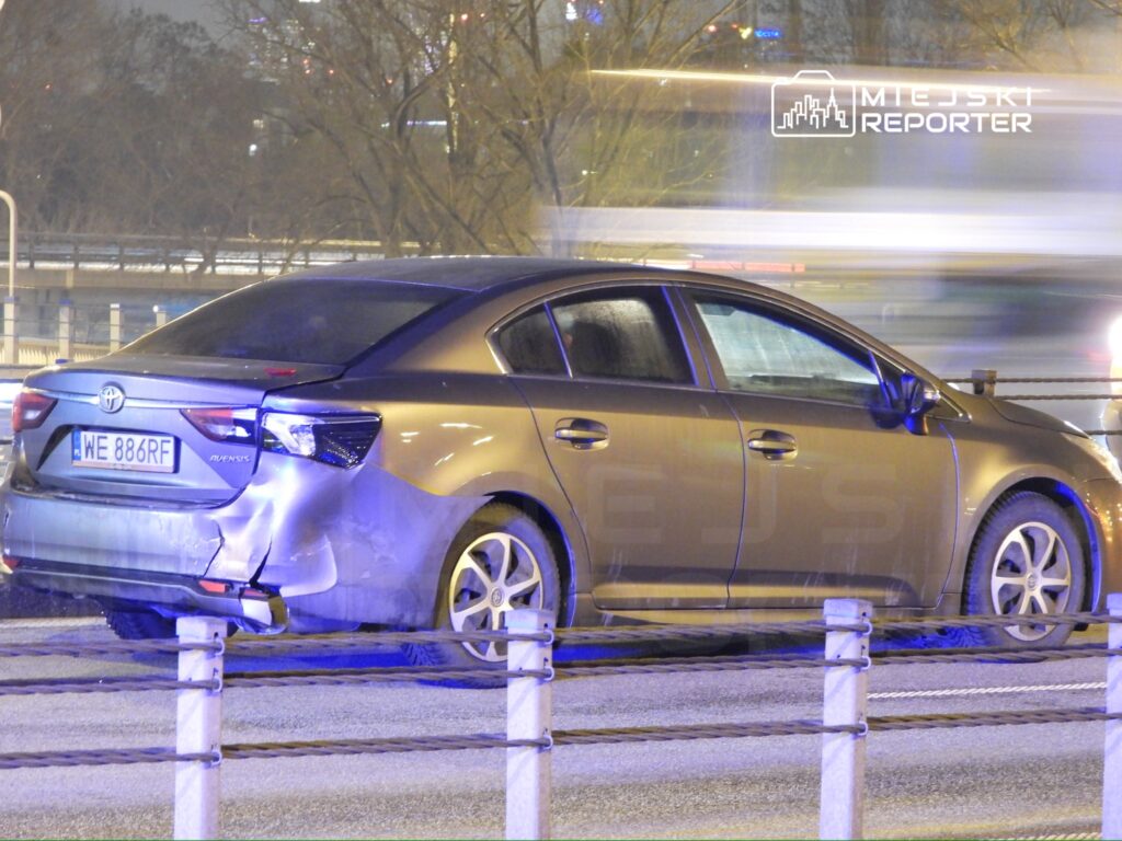 Uszkodzony samochód osobowy zaparkowany na poboczu drogi, w tle widać rozmyte światła przejeżdżających pojazdów.