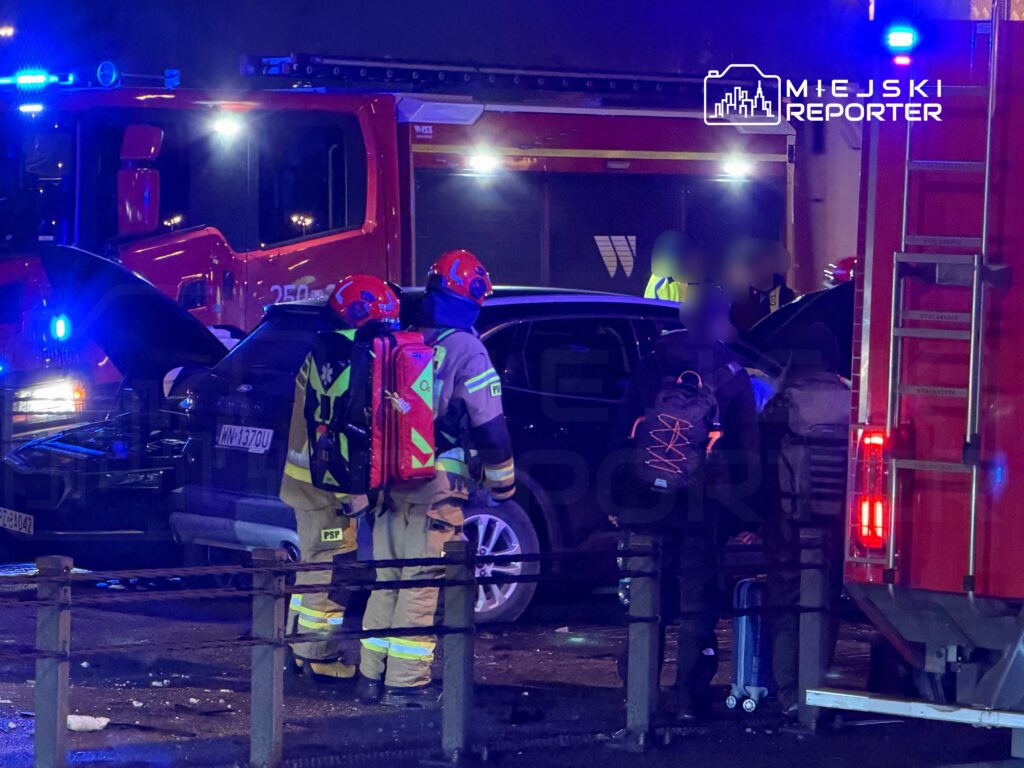 Ratownicy medyczni sprawdzają poszkodowanego w czarnym samochodzie po wypadku, obok stoi wóz strażacki.