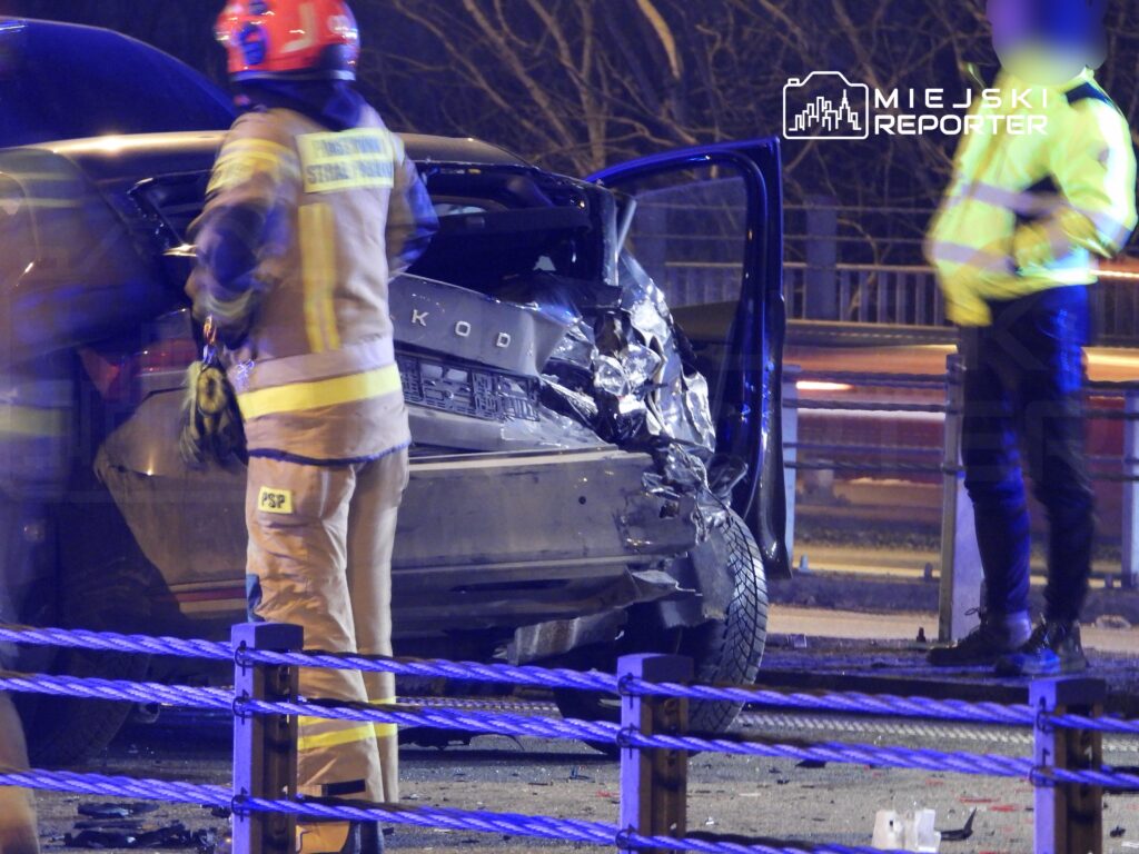 Strażak w mundurze sprawdza uszkodzony samochód osobowy na drodze, obok stoi policjant w odblaskowej kurtce.