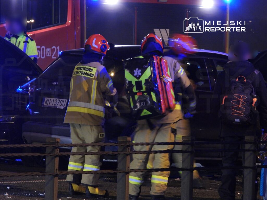 Strażacy w mundurach interweniują przy zderzonym samochodzie w nocnym otoczeniu miejskim.