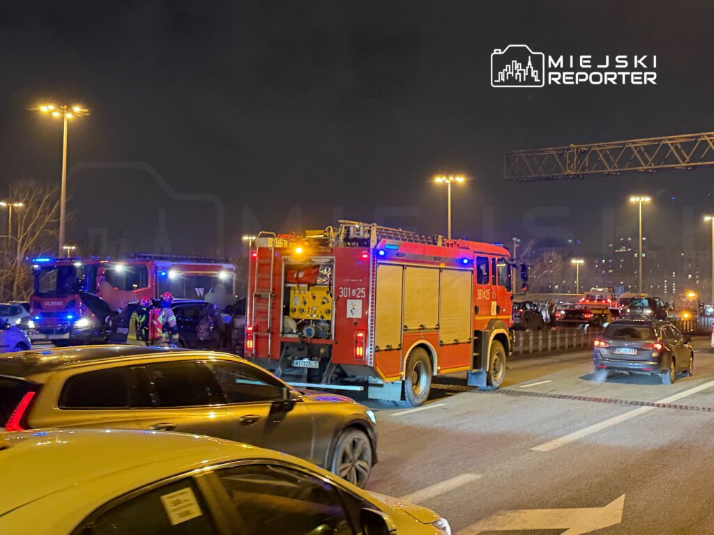 Strażacy w hełmach rozmawiają obok wozu strażackiego na zatłoczonej drodze w nocy. W tle widać inne pojazdy.