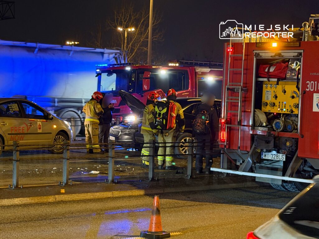 Grupa strażaków w mundurach interweniuje przy uszkodzonym samochodzie na drodze, obok stoi wóz strażacki.
