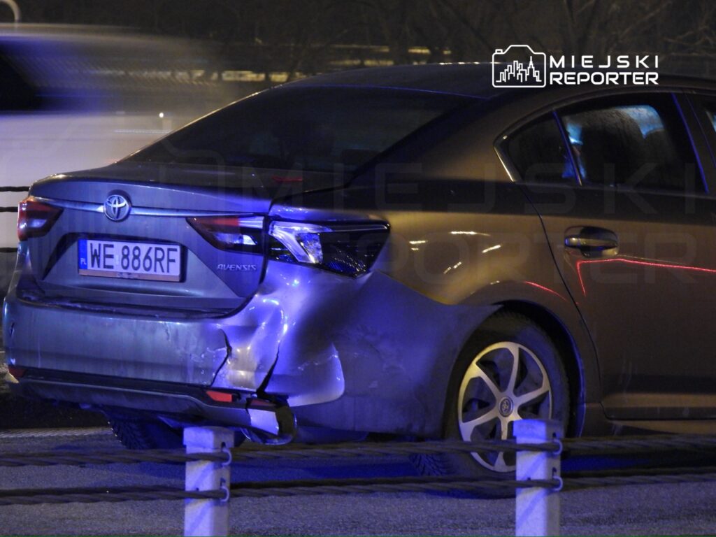 Uszkodzony samochód marki Toyota Avensis zaparkowany w nocy na ulicy, z widocznymi śladami kolizji.