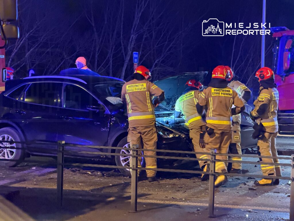 Czterech strażaków w mundurach z hełmami bada uszkodzony samochód osobowy po wypadku drogowym w nocy.