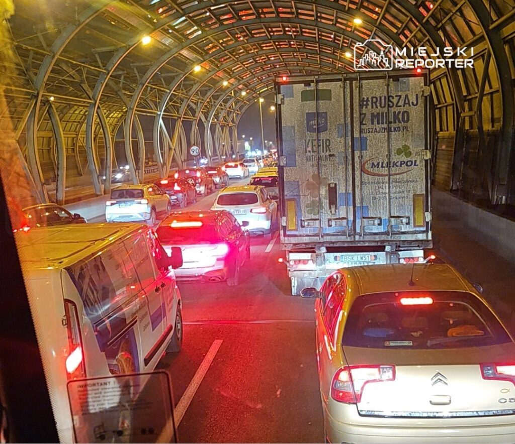 Korki drogowe w tunelu, z różnymi samochodami stojącymi w zatorze, nocą przy oświetleniu ulicznym.