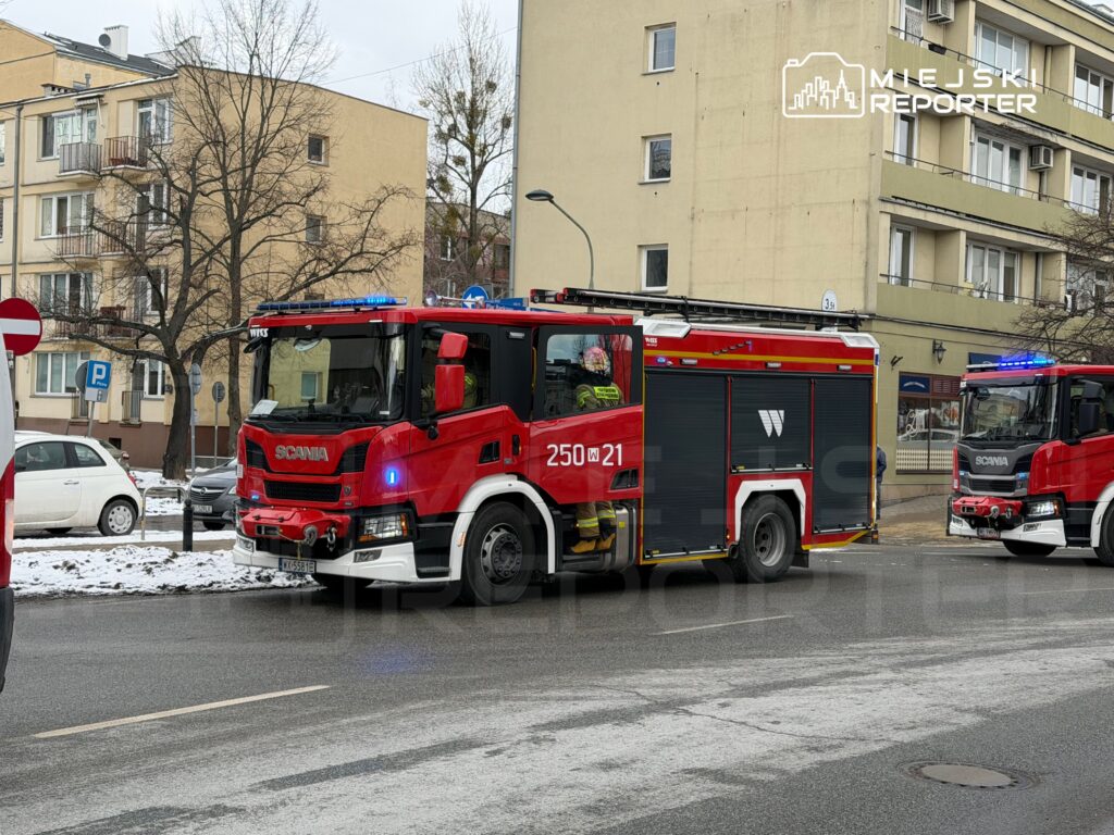 Czerwony wóz strażacki z włączonymi sygnalizatorami zatrzymany na ulicy w mieście, obok budynków mieszkalnych.
