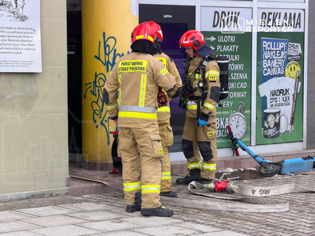 Grupa strażaków w mundurach omawia sytuację przed budynkiem z graffiti, obok sprzętu gaśniczego.
