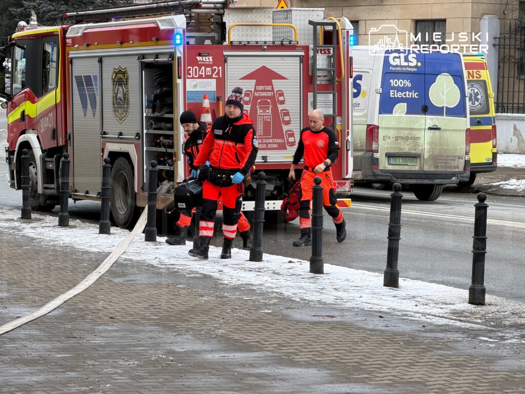 Ratownicy w czerwonych mundurach wynoszą sprzęt z wozu strażackiego na ośnieżonym chodniku.