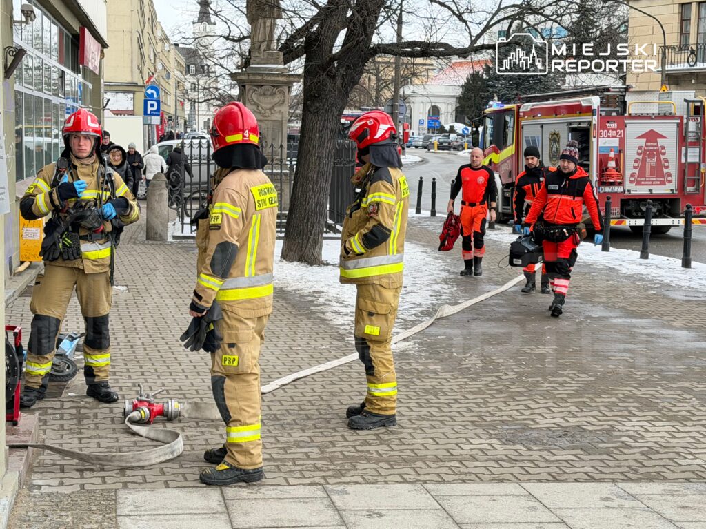 Grupa strażaków w mundurach omawia sytuację na chodniku, obok węża strażackiego i wozu bojowego.