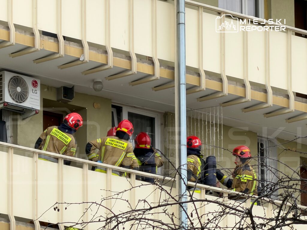 Grupa strażaków w hełmach omawia działania ratunkowe na balkonie budynku mieszkalnego.