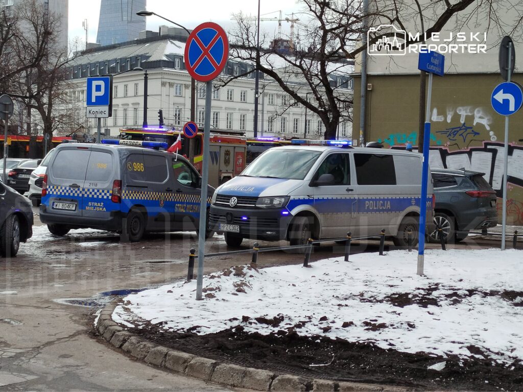 Na skrzyżowaniu ulic stoją pojazdy Straży Miejskiej i Policji, a w tle widoczny jest zasypany śniegiem chodnik.
