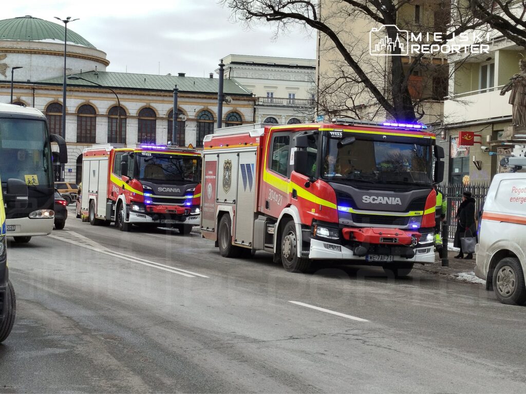 Dwa wozy strażackie marki Scania z włączonymi sygnałami świetlnymi przejeżdżają obok nowoczesnego budynku.