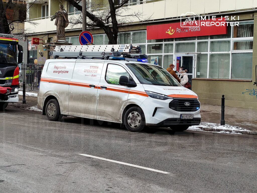 Pojazd pogotowia energetycznego zaparkowany przed budynkiem poczty, obok mężczyzny z psem.