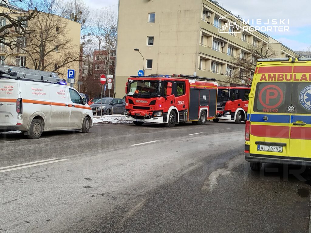 Na ulicy parkują dwa pojazdy służbowe: jeden z logo Pogotowia Energetycznego oraz ambulans, obok stoi wóz strażacki.
