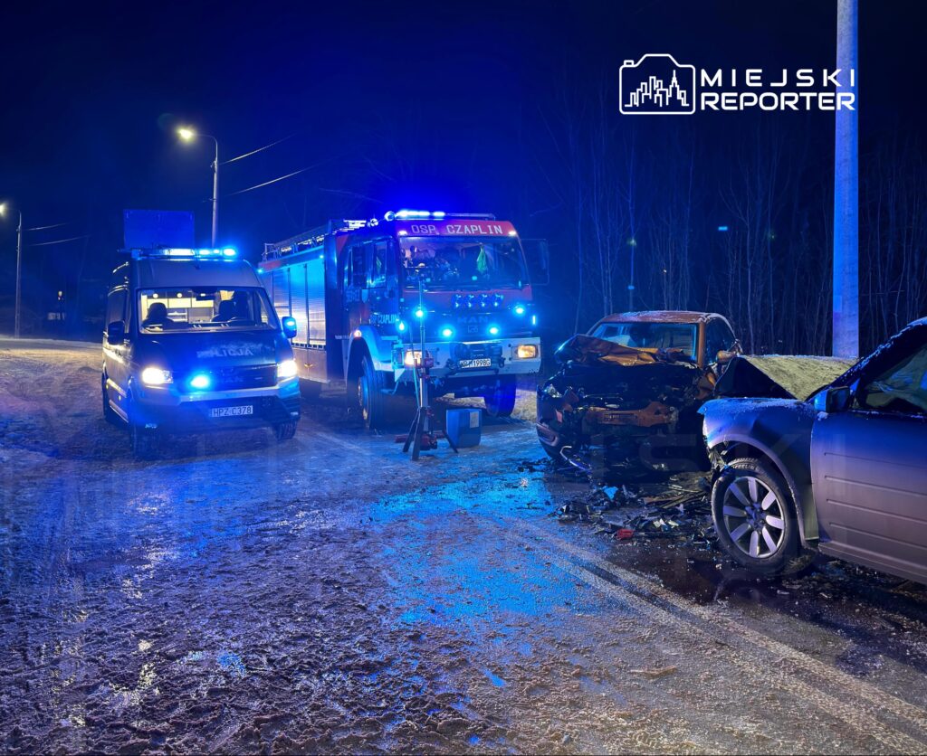 Na oświetlonej nocą drodze stoi nieoznakowany radiowóz oraz wóz strażacki, obok widoczny wypadek dwóch uszkodzonych samochodów.