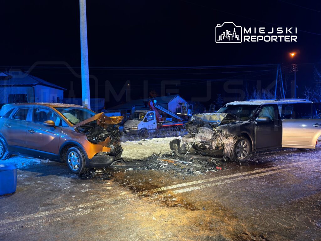 Dwa zniszczone pojazdy po kolizji na oświetlonej ulicy, obok widoczne ślady wypadku i uszkodzone mienie.