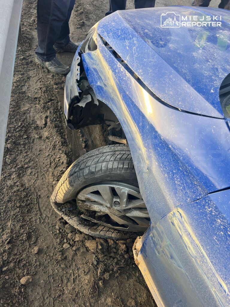 Zniszczony niebieski samochód osobowy z uszkodzoną przednią częścią i przebitą oponą na ziemistej nawierzchni.