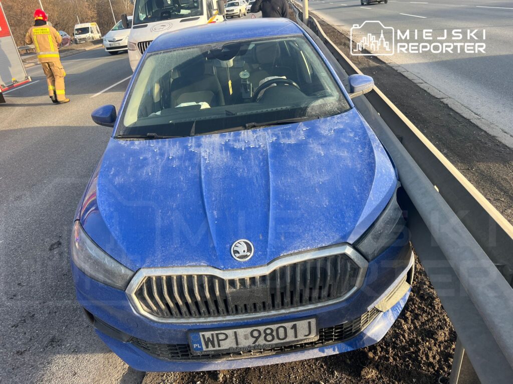 Niebieski samochód osobowy zaparkowany przy barierce na drodze, obok strażak w mundurze.