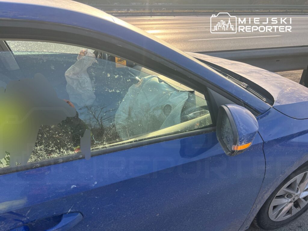 Zniszczony niebieski samochód osobowy z wystrzeloną poduszką powietrzną w kabinie, zaparkowany przy drodze.