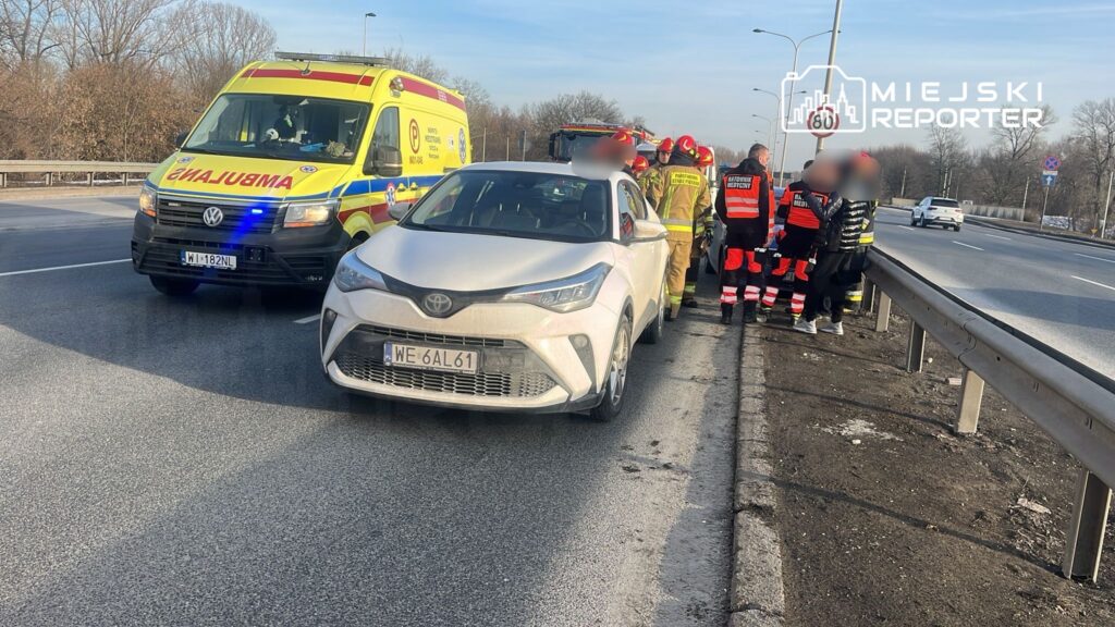 Na drodze stoi biały samochód osobowy, obok grupka ratowników medycznych i strażaków, a w tle widoczna karetka.