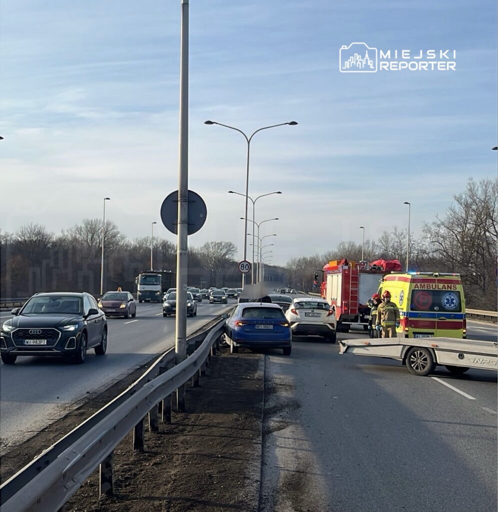 Na pasie drogowym stoi niebieski samochód osobowy, a obok niego pracują strażacy i zespół ratownictwa medycznego. W tle widać ruchliwe pojazdy oraz znaki drogowe.