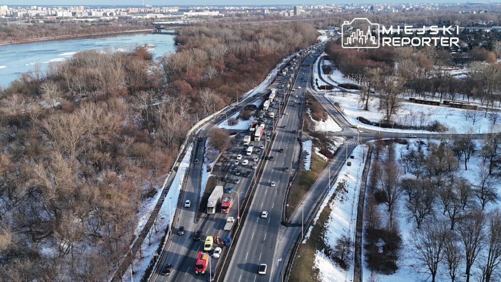 Ruch drogowy na zatłoczonej trasie, z widokiem na rzekę i osiedla w tle, w zimowej scenerii.