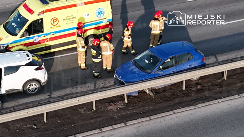 Grupa strażaków w hełmach rozmawia obok niebieskiego samochodu na drodze, podczas gdy ambulans stoi w tle.
