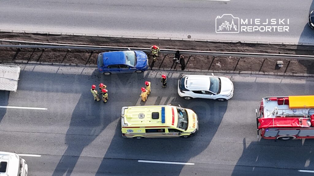 Grupa strażaków w hełmach omawia sytuację obok niebieskiego samochodu na poboczu drogi, obok karetki i wozu strażackiego.