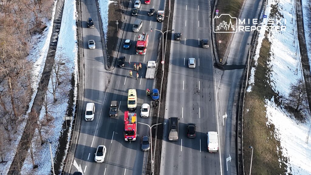 Na drodze widoczne są pojazdy, w tym karetka i wóz strażacki, oraz ratownicy udzielający pomocy przy stłuczce.