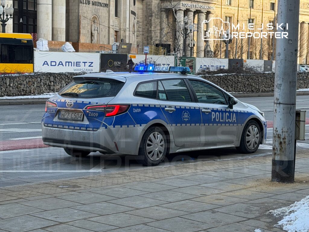 Policyjny samochód osobowy z niebieskimi światłami zaparkowany na ulicy w pobliżu budynków.