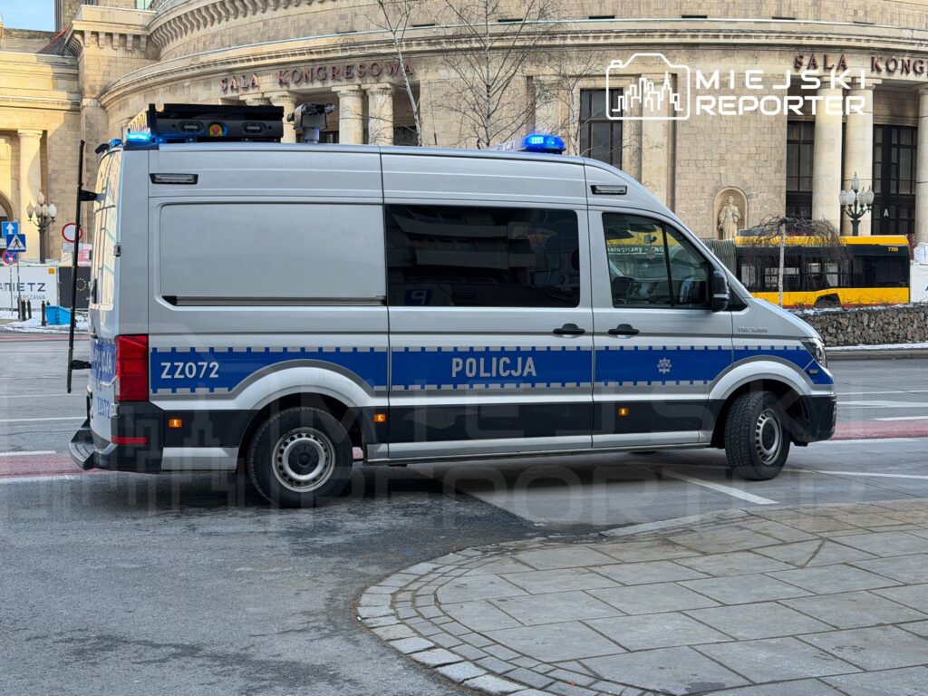 Policyjny van zaparkowany na ulicy przed Salą Kongresową, z włączonymi niebieskimi światłami.