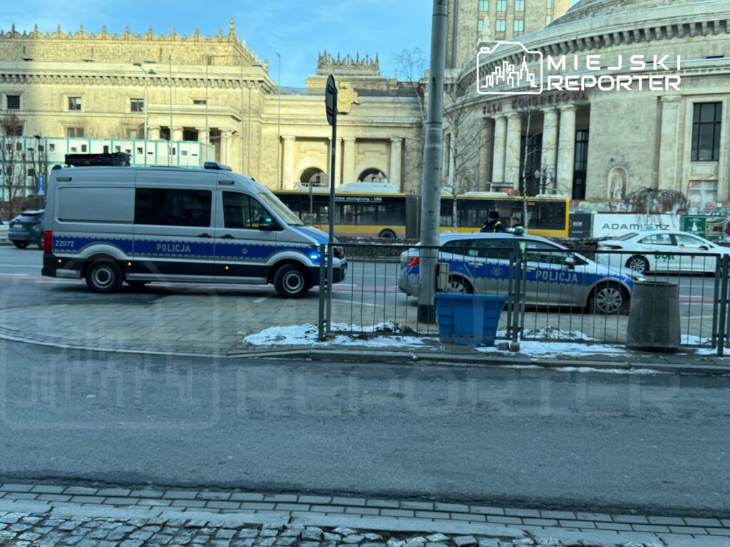 Policyjny van oraz radiowóz zatrzymane na ulicy w otoczeniu architektury miejskiej i komunikacji publicznej.